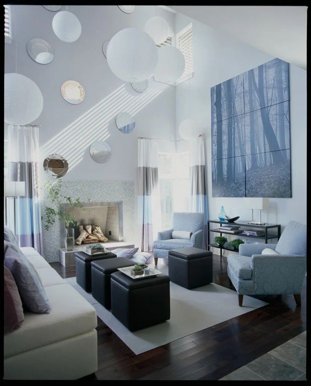 Bright, high-ceilinged contemporary living room with white walls, dark timber flooring, a large pale area rug, and multiple upholstered sofas and armchairs arranged around black ottomans and a central coffee table. A stone fireplace, sheer curtains, circular wall mirrors, spherical pendant lights, and a large blue-toned forest artwork create a cool, refined interior.