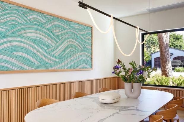 Bright contemporary dining area with a large white marble-look dining table, curved timber chairs, and a timber slatted half-height wall treatment. A large turquoise abstract artwork hangs on the white wall, while a sculptural linear pendant light spans the ceiling. Full-height corner glazing provides views to a landscaped exterior.