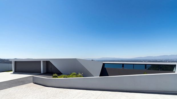 A low-profile contemporary building viewed from an elevated exterior terrace or rooftop. The structure has crisp geometric forms, smooth white and dark grey rendered surfaces, a long horizontal strip of blue-tinted glazing, and a broad parapet wall. A distant urban landscape and mountain range are visible beyond the building under a clear blue sky.