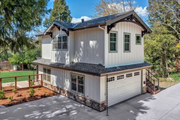A compact two-storey detached outbuilding or residence with pale vertical board-and-batten siding, dark shingle gable roof, decorative timber gable brackets, stone-clad lower walls, and white-framed windows. A garage occupies the lower level, with a small stair access to the upper floor. The building sits beside a landscaped yard and concrete driveway, surrounded by mature trees.