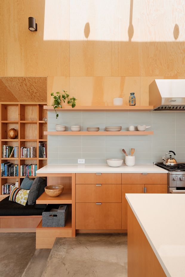 A compact, warm-toned kitchen with extensive natural timber cabinetry and wall panelling. The space includes open shelving with books and kitchenware, a pale tiled splashback, white benchtops, integrated shelving and bench seating, a freestanding stainless-steel range cooker, and a small plant. Concrete-look flooring and restrained furnishings create a practical, modern interior with strong timber detailing.