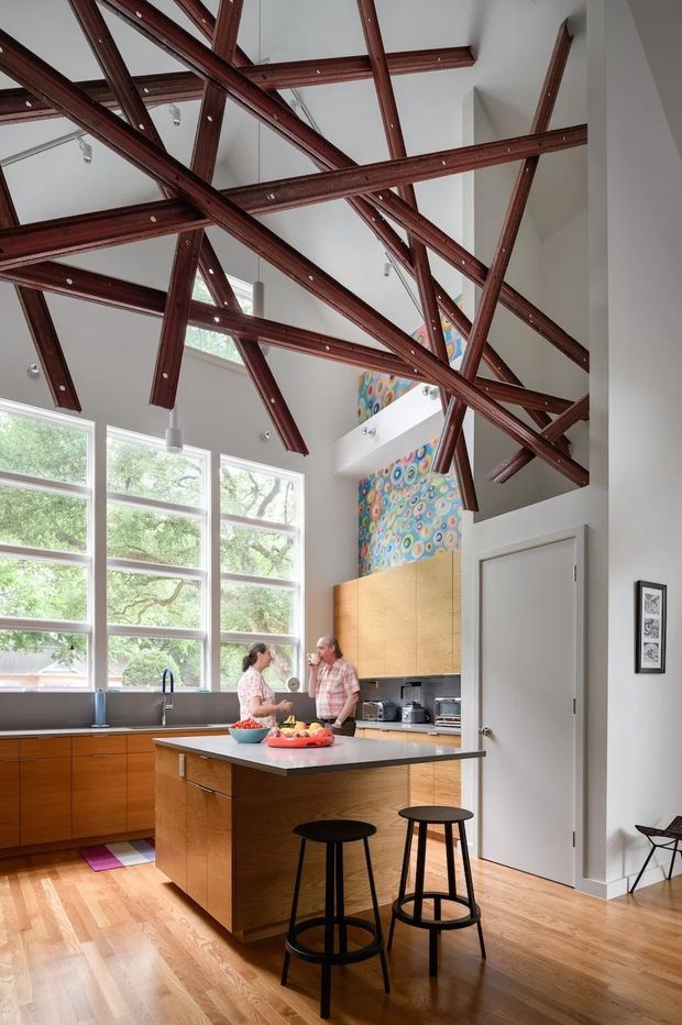 A bright, double-height contemporary kitchen with large multi-pane windows, warm timber cabinetry, a central island, and black bar stools. The standout architectural feature is an exposed network of dark timber structural beams crossing the ceiling and upper volume. White walls, timber flooring, pendant lights, and colourful artwork complete the space.