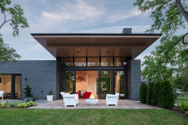 Contemporary single-storey house facade with dark horizontal brick walls, extensive floor-to-ceiling glazing, and a broad flat roof with a warm timber-lined soffit. A timber deck creates an outdoor seating area with white lounge chairs and a red accent chair, opening onto a landscaped lawn with mature trees and hedges.