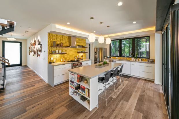 Bright, spacious contemporary kitchen with a large central island and integrated open shelving, white flat-panel cabinetry, and a vivid yellow patterned tile backsplash. The kitchen has warm wood-look flooring, dark-framed windows, pendant lighting, and an adjoining hallway visible to the left.