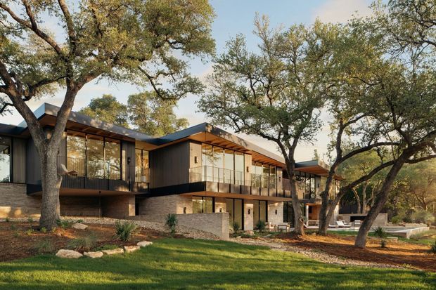 Exterior view of a contemporary multi-level residence set among mature trees and landscaped grounds. The home features strong horizontal and cantilevered volumes, dark metal cladding, extensive floor-to-ceiling glazing, projecting balconies with slender metal balustrades, timber-toned soffits, and a low stone retaining wall. The setting appears wooded and suburban or rural, with warm late-day light.