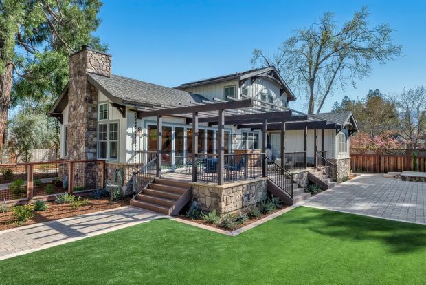 Exterior view of a two-storey suburban home with a gabled roof, prominent stone chimney and stone-clad lower walls. The house features a covered rear porch or terrace with dark timber framing, screened or glazed sections, steps, and metal railings. Paved paths and a broad paved courtyard surround the home, with landscaped garden beds and lawn.