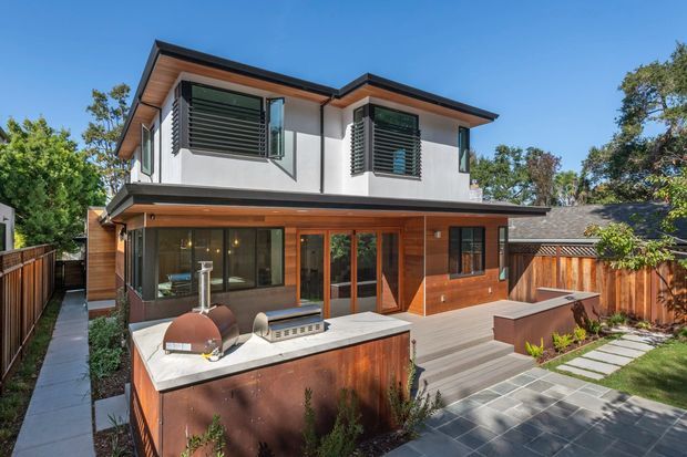 Exterior view of a modern two-storey residence with a low-pitched asymmetrical roof, white rendered upper level, warm timber-clad ground floor, dark-framed glazing, and external louvred window screens. A paved courtyard and timber deck surround an outdoor kitchen with built-in cabinetry and a pizza oven, enclosed by timber fencing and landscaping.