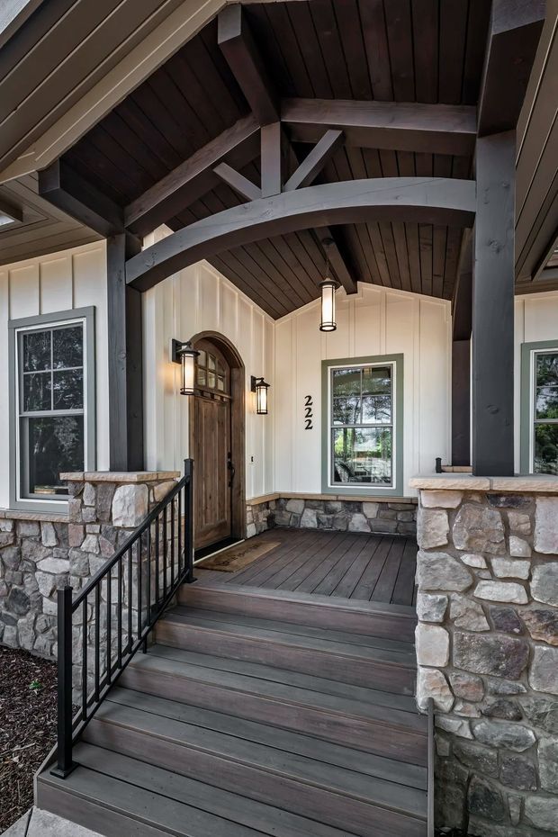 A covered residential front porch viewed from the steps, featuring dark timber decking and exposed timber beams beneath a vaulted gable roof. The entry has a natural timber arched door, white vertical wall cladding, dark-framed windows, black lantern wall lights, black metal railings, and substantial irregular natural-stone masonry columns and base walls.