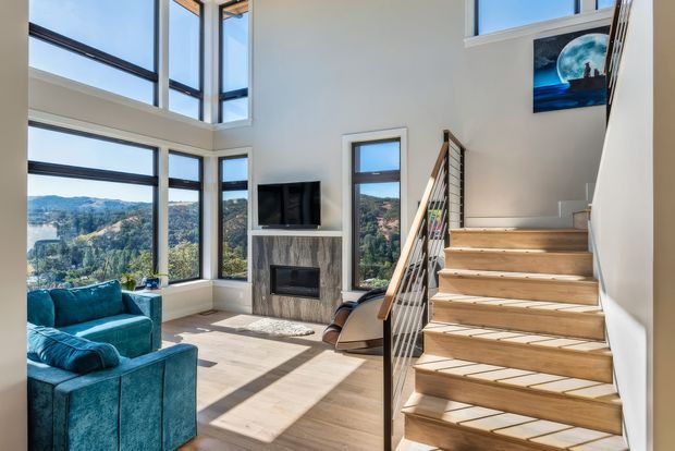 Bright, double-height contemporary living room with expansive floor-to-ceiling corner glazing overlooking a wooded hillside. The room includes a teal sectional sofa, a built-in fireplace with stone-look surround and television, light timber flooring, and an open timber staircase with dark metal balustrade leading to an upper level. Strong natural light and dramatic vertical glazing create an airy, elevated interior.