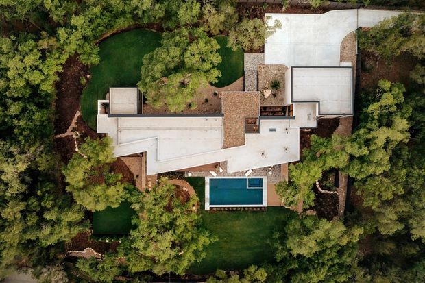 Aerial top-down view of a contemporary multi-wing residence surrounded by dense landscaped greenery. The property has several flat and low-pitched roof sections, pale concrete or rendered roof surfaces, a brown tiled roof section, large paved outdoor areas, and a rectangular swimming pool adjacent to the house. Curving garden paths and lawns weave through the mature vegetation.