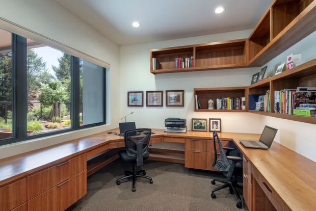 A bright, custom-built home office with a large corner workstation, extensive warm timber cabinetry, open shelving filled with books and framed artwork, and two ergonomic office chairs. A broad window provides views onto greenery, while recessed ceiling lights evenly illuminate the room. The space has a practical, refined, built-in study design.