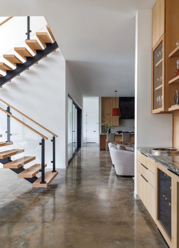 A bright, contemporary open-plan interior viewed from a polished concrete hallway. The foreground features a sculptural open-riser staircase with pale timber treads, black steel stringers and posts, and clear glass balustrades. To the right is built-in timber cabinetry with glass-front display storage and a stone benchtop, while the space opens toward a dining and living area with timber joinery, upholstered seating, and a bold red pendant light. White walls, extensive glazing, and reflective flooring create an airy, modern atmosphere.