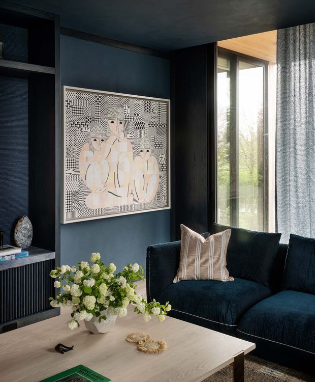 A moody, refined living room with deep blue-black walls, ceiling and built-in shelving. A large graphic artwork is mounted above a dark fluted cabinet, while a deep navy sofa with a striped neutral cushion faces a light timber coffee table. Floor-to-ceiling glazing with textured curtains brings in natural light, and a sculptural floral arrangement provides a soft organic contrast.