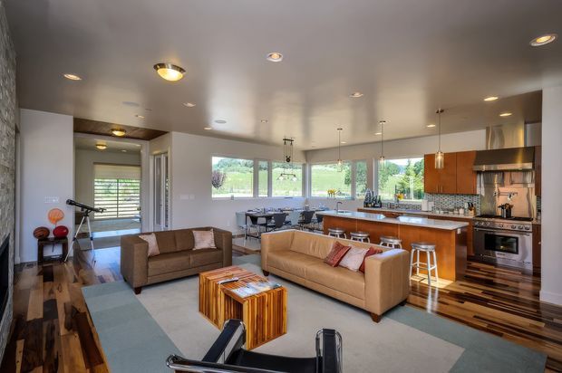 A spacious open-plan living, dining, and kitchen area with a contemporary fit-out. The room features warm timber flooring, a large pale area rug, two tan sofas, a timber coffee table, a dining table near the windows, and a long kitchen island with bar stools. The kitchen has warm wood cabinetry, light benchtops, stainless steel appliances, pendant lighting, and broad windows overlooking greenery. Recessed ceiling lights and a dark stone-textured feature wall add contrast.