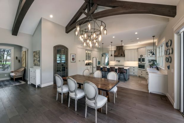 A spacious open-plan dining and kitchen interior with a large dark timber dining table, upholstered dining chairs, white cabinetry, island seating, and integrated appliances. The room features exposed dark timber ceiling beams, a vaulted ceiling, arched openings and windows, dark wood-look flooring, pendant lights, and a prominent multi-light chandelier over the dining table. A small sitting area is visible to the left.