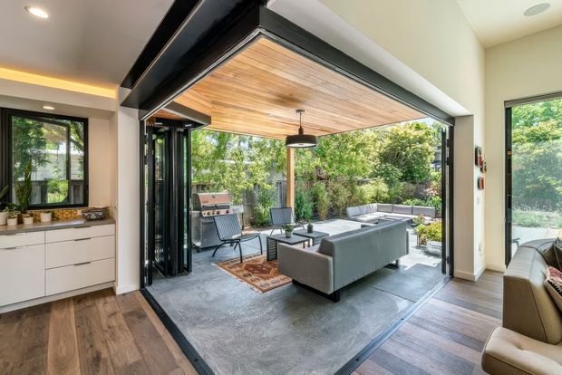 A bright contemporary open-plan living area opening through fully retractable black-framed glass doors to a covered outdoor entertaining space. The interior has warm timber flooring, white built-in cabinetry, and neutral upholstered seating. Outside, a timber-lined ceiling, outdoor sofa, dining setting, and barbecue create a seamless indoor-outdoor living zone surrounded by greenery.