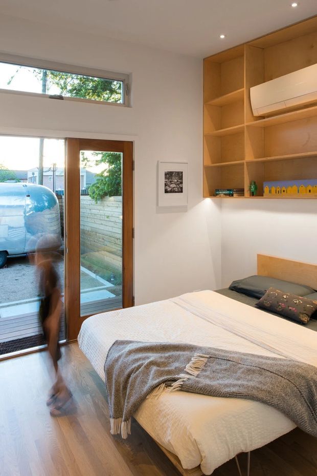 A compact contemporary bedroom with a double bed, white bedding, and a textured grey throw. The room features warm timber flooring and a full-height built-in timber shelving and storage wall with an integrated split-system air conditioner. A timber-framed glazed door and high horizontal window provide daylight and views to the exterior. Recessed ceiling lights and minimal decor create a clean, warm atmosphere.