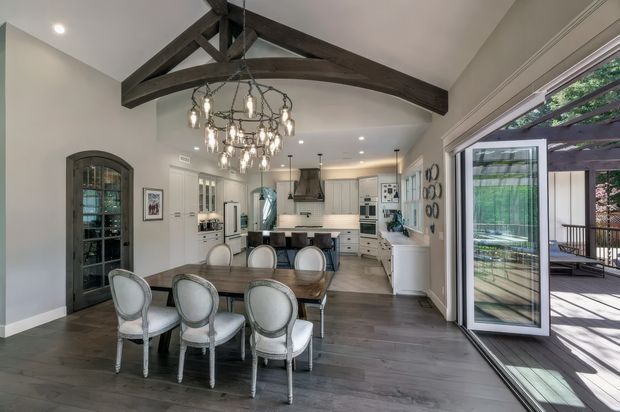 A bright open-plan dining and kitchen interior with a long timber dining table, upholstered dining chairs, white cabinetry, a central island, and a large open glass door leading to an outdoor deck. The room features a vaulted ceiling with prominent dark exposed timber beams and a large ornate chandelier, blending traditional architectural details with a refined contemporary fit-out.