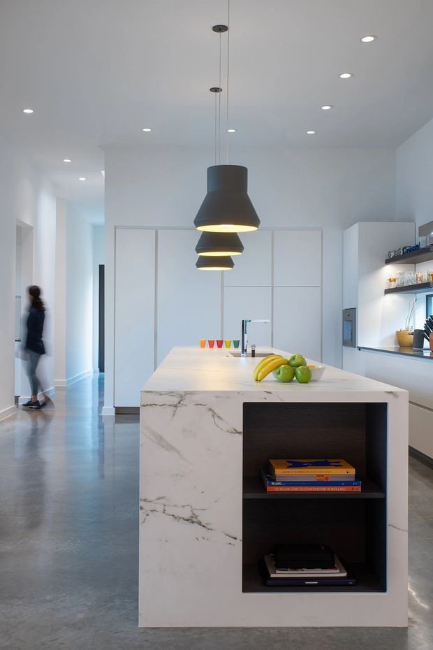 A bright, minimalist contemporary kitchen viewed from the main living circulation area. The room features a large white marble island with an integrated dark open shelving niche, a pale engineered or natural stone benchtop, concealed white cabinetry, a reflective grey polished concrete floor, recessed ceiling lights, and three black sculptural pendant lights. A rear wall includes open display shelving and a window or serving opening. The kitchen connects to a hallway, with a blurred person at the left edge.