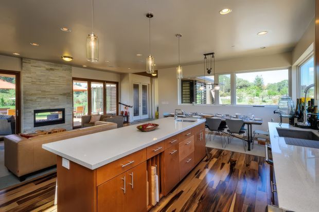 Open-plan contemporary kitchen and living area with a large central island, warm timber cabinetry, white stone benchtops, polished dark timber flooring, pendant lighting, and expansive windows. A lounge with a built-in fireplace is visible beyond the kitchen, with a dining area beside the glazing.