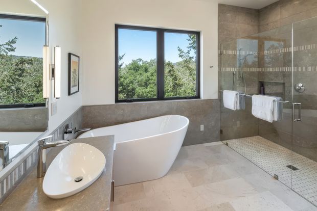 A bright, spacious contemporary bathroom featuring a freestanding white bathtub, sculptural white vessel basin on a stone vanity, large dark-framed windows, and a walk-in glass shower with a tiled feature wall. Natural greenery is visible outside the windows, while the bathroom is finished in warm beige and grey stone tones.