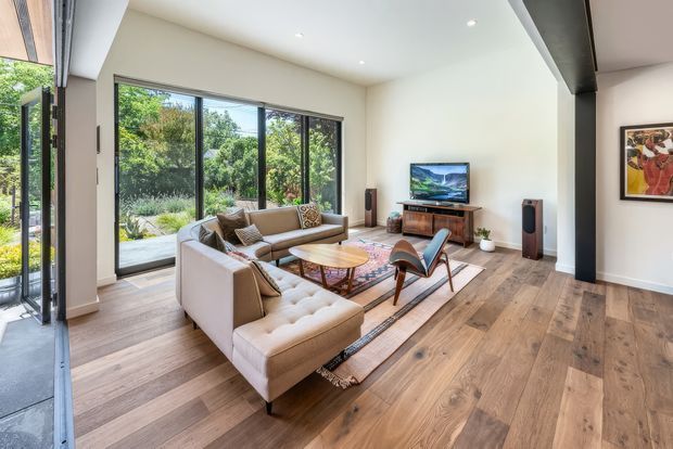 Bright contemporary living room with warm wide-plank timber flooring, a large cream sectional sofa, mid-century style lounge chair, round timber coffee table, patterned rug, and a compact media console with television and speakers. Full-height black-framed glazing opens toward a landscaped garden, while a prominent dark structural beam and recessed ceiling lights add architectural definition.
