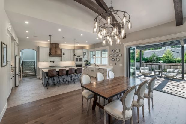 Bright, spacious open-plan kitchen and dining area with a large island, white cabinetry, stainless-steel appliances, and pendant lights. A wood dining table with upholstered chairs sits in the foreground beneath an ornate chandelier. Exposed dark timber ceiling beams, light tiled flooring in the kitchen, and wide glazed doors opening to a timber outdoor deck are visible.