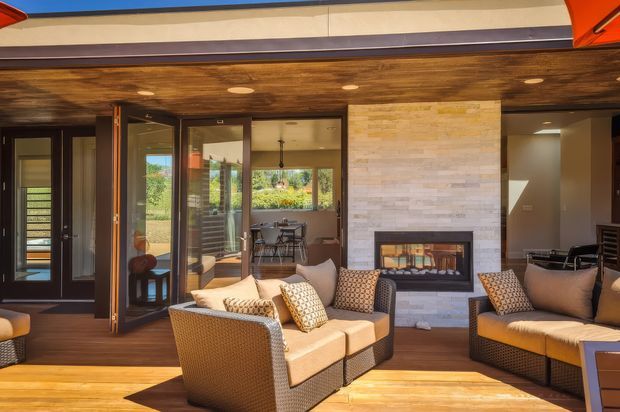 A warm, covered outdoor living area with timber decking, woven lounge seating, a built-in fireplace set into a light stone feature wall, and a timber-lined ceiling. Large glazed folding or sliding doors connect the space to an interior dining area and garden beyond. The setting is bright and relaxed, with strong indoor-outdoor connection.