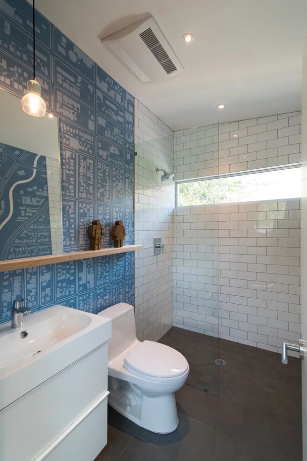 A compact contemporary bathroom with a white wall-mounted vanity and basin, toilet, frameless glass shower enclosure, dark floor tiles, and white subway wall tiles. One wall features a striking blue graphic/map-patterned tile or wallpaper-like surface with a slim timber display shelf. A horizontal clerestory-style window brings in daylight, while pendant and recessed lighting provide illumination.