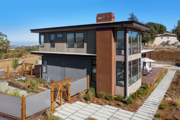 Exterior view of a contemporary multi-level residence on a landscaped sloping site. The house features dark vertical cladding, a prominent rust-coloured metal-clad vertical volume, broad floor-to-ceiling glazing, a low-pitched flat roof, and a rooftop chimney or vent. Paved paths, timber garden structures, and planted terraces surround the building, with distant coastal scenery visible beyond.