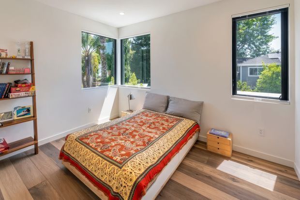 Bright bedroom with a low bed dressed in an ornate red, orange and gold patterned coverlet, light neutral walls, warm wood flooring, a simple wooden bookshelf, and a small bedside drawer unit. Large corner windows and a separate window provide abundant natural light and views of greenery and a neighbouring home. The room has a simple, contemporary shell with eclectic patterned textiles.