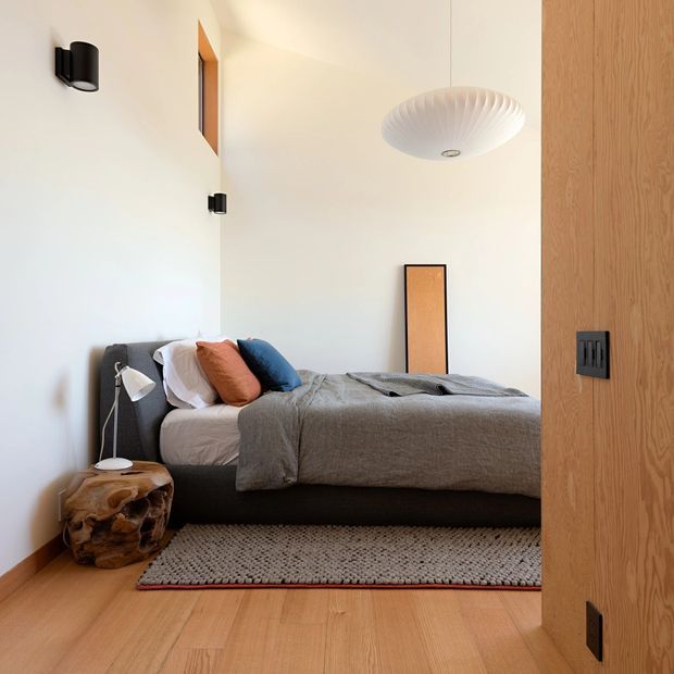 A calm, compact bedroom viewed through an open timber door. The room features a low dark upholstered bed with grey linen bedding, white, rust and blue cushions, a textured neutral rug, warm timber flooring and matching timber wall or door detailing. White walls, black wall lights, a sculptural pendant and a rustic organic bedside stool complete the understated modern interior.