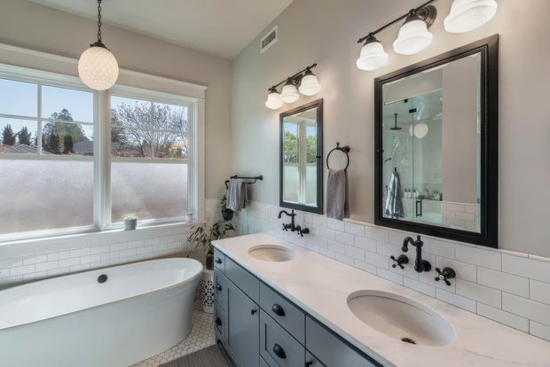 Bright, refined bathroom with a freestanding white bathtub beside frosted and clear windows, a long double-sink vanity with dark blue-grey cabinetry and white countertop, white subway-tile splashback, black-framed mirrors, and traditional black wall-mounted fittings. The large mirror reflects a glass shower enclosure. Natural daylight creates a clean, inviting atmosphere.