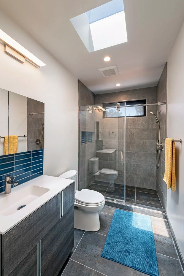 A modern bathroom with a floating dark wood-grain vanity, white integrated basin, large wall mirror, vivid blue tiled splashback, and a glass-enclosed walk-in shower. The shower and floor use large grey stone-look tiles, with a skylight, recessed ceiling lights, a horizontal clerestory-style window, and a white toilet. Yellow towels and a blue bath mat add colour accents.