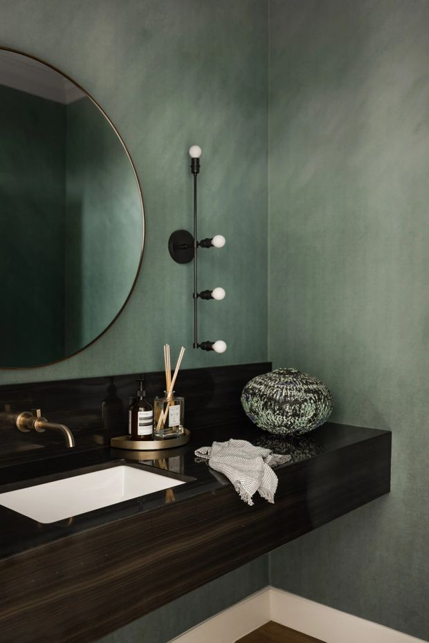 A moody, refined bathroom vanity featuring deep dark timber joinery with an integrated white rectangular basin, brass wall-mounted tapware, dark splashback, and textured green painted walls. A large round mirror and vertical globe wall sconce provide strong decorative focal points, while a small vase, toiletries, and folded hand towel sit on the vanity.