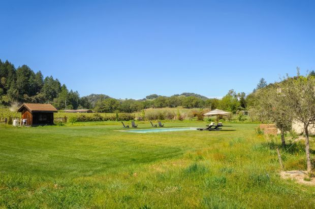 A spacious rural landscaped property with a broad green lawn, a small timber pavilion or shed at left, a narrow swimming pool with loungers in the middle distance, and a parasol-covered seating area. Mature trees and open vegetation surround the grounds under a clear blue sky.