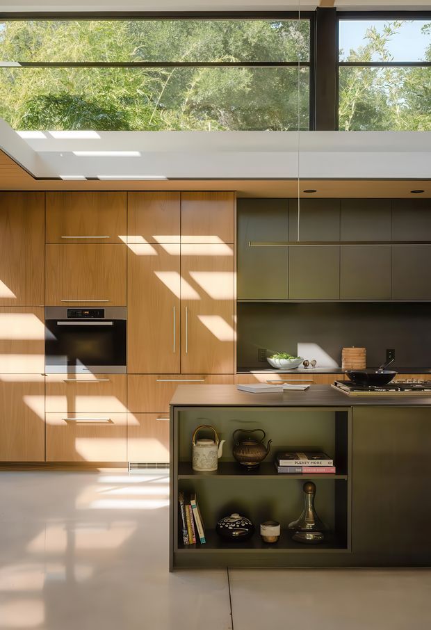 A contemporary kitchen with warm timber cabinetry, integrated stainless-steel appliances, a dark reflective backsplash, and a central island with open display shelving. High clerestory-style glazing brings in abundant natural light and views of leafy trees, while strong sunlight creates geometric shadows across the cabinetry and polished floor. The kitchen has a refined, gallery-like appearance with muted olive and charcoal tones contrasted by honey-coloured timber.