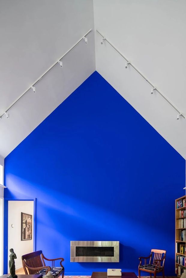 A dramatic double-height living room dominated by a vivid cobalt-blue feature wall rising to a pitched ceiling. White walls and exposed track lighting frame the space, with two timber lounge chairs, a low coffee table, a wall-mounted fireplace with a metallic surround, and built-in bookshelves visible at the sides. A doorway opens to an adjacent room on the left.
