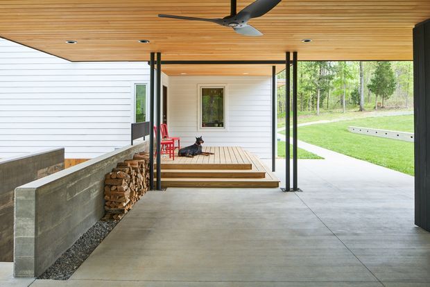 A covered outdoor living and circulation area framed by slim dark posts, with a warm timber-lined ceiling, smooth concrete floor, white horizontal cladding, and a raised timber porch beyond. A built-in concrete-edged wood storage feature sits along the left, while the open side looks onto a landscaped lawn and path. A ceiling fan and recessed lighting provide functional detailing.
