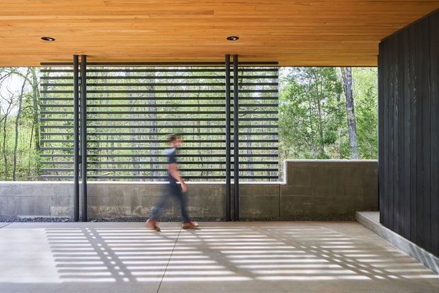 A covered modern outdoor passage or terrace with a warm timber-lined ceiling, polished concrete floor, dark vertical wall cladding, and a horizontal louvred screen framing views into a wooded landscape. Strong striped shadows fall across the floor, with a blurred person walking through the space. The composition emphasises indoor-outdoor connection, filtered light, and restrained contemporary detailing.