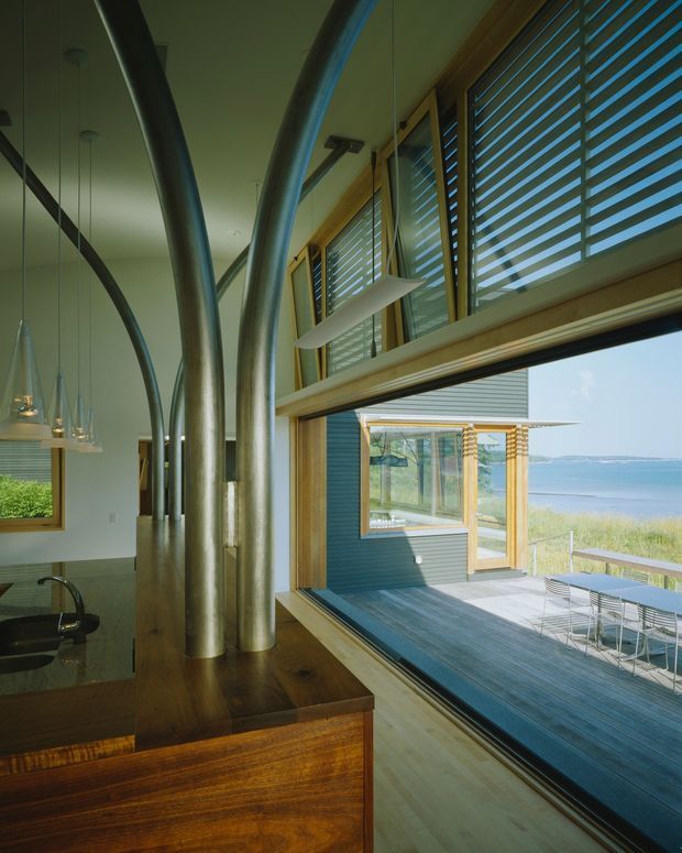 Interior view of a contemporary coastal living and dining space opening onto a timber deck, with expansive ocean-facing glazing, horizontal louvres, warm timber joinery, and sculptural curved metal columns. A dining area and kitchen counter are visible inside, while the deck and ocean are seen through the large open glazed wall.