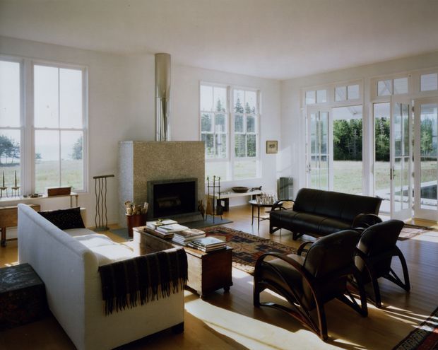 Bright, spacious living room with large multi-pane windows and French doors overlooking a grassy landscape. The room features polished timber flooring, a substantial stone fireplace with a dark hearth, a white sofa, black leather seating, a patterned area rug, and mid-century-style furniture arranged around a central coffee table.
