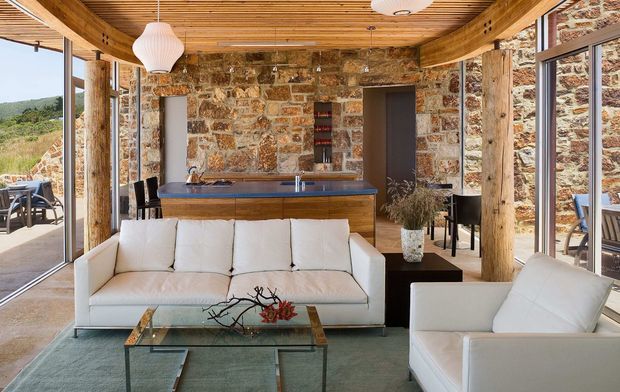 Open-plan kitchen and living area with a white sofa and armchair, compact island, exposed rustic stone walls, substantial timber posts and beams, and a warm slatted timber ceiling. Full-height glazing opens toward an outdoor terrace and landscape, while contemporary furniture and pendant lighting contrast with the natural structural materials.