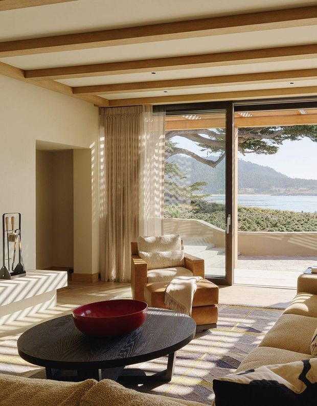 Warm, refined living room with a low black oval coffee table, sculptural red bowl, upholstered armchairs and a large sectional sofa. Exposed timber ceiling beams contrast with a light plaster ceiling and walls. Floor-to-ceiling glazed doors open to a terrace, revealing a coastal bay and wooded hills beyond; sunlight creates strong linear shadows through the glazing and curtains.
