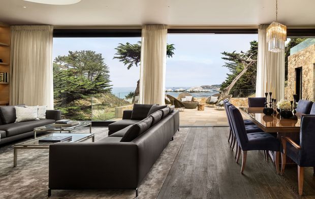 A refined open-plan living and dining room with expansive floor-to-ceiling glazing overlooking a coastal landscape and ocean. The room features dark upholstered sofas, a large glass coffee table, a timber dining table with upholstered chairs, full-height curtains, wide-plank timber flooring, and a textured stone feature wall. A terrace and outdoor seating are visible beyond the glazing.