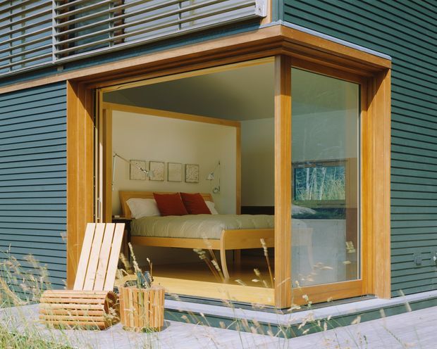 Exterior view of a compact timber-framed bedroom extension or glazed bedroom pod, set within dark horizontal siding. Large corner glazing and an open glass door reveal a simple bed, pale walls, timber flooring, and warm wood trim inside. A small outdoor deck with a timber chair and planter is visible in the foreground.