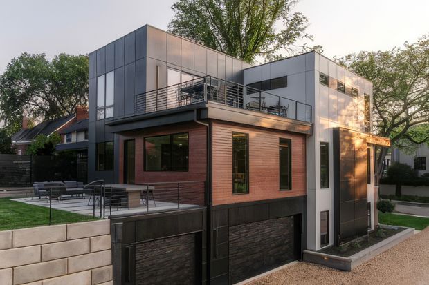 Exterior view of a contemporary multi-level urban residence with a boxy flat-roof form, dark metal-clad upper volumes, warm horizontal timber cladding, extensive glazing, rooftop terrace, and a dark stone-clad lower level. The home sits within a landscaped suburban setting and includes a paved outdoor area and perimeter retaining wall.