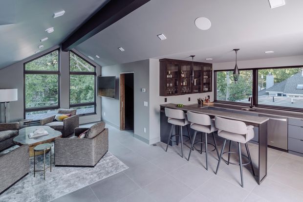 Open-plan contemporary kitchen and living area with a vaulted ceiling and exposed dark structural beam. The kitchen features a long island with a dark stone-look benchtop, integrated cabinetry, a glass-front display cabinet, and four upholstered bar stools. Large black-framed windows provide abundant natural light and views of trees and neighbouring homes. The adjoining living zone contains grey upholstered armchairs and sofas around a low coffee table on a large rug.