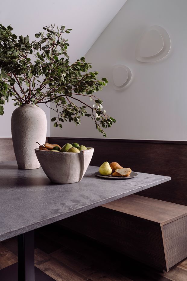 A refined contemporary dining area featuring a dark timber built-in banquette or sideboard beneath a thick grey stone dining table. A sculptural ceramic vase with leafy branches and matching ceramic fruit bowls add organic forms, while minimalist raised wall details provide subtle decoration. The palette is muted grey, dark brown and natural wood, creating a sophisticated, moody atmosphere.