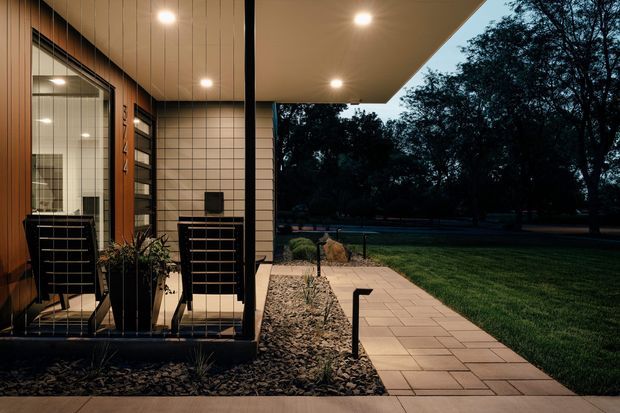 Dusk view of a modern residential entry and covered porch. The facade combines vertical timber cladding, light rectangular masonry or tiled wall panels, dark metal-framed glazing, and a broad flat-roof canopy. A paved walkway crosses a landscaped gravel bed with low planting and discreet pathway lighting, opening toward a lawn and mature trees.